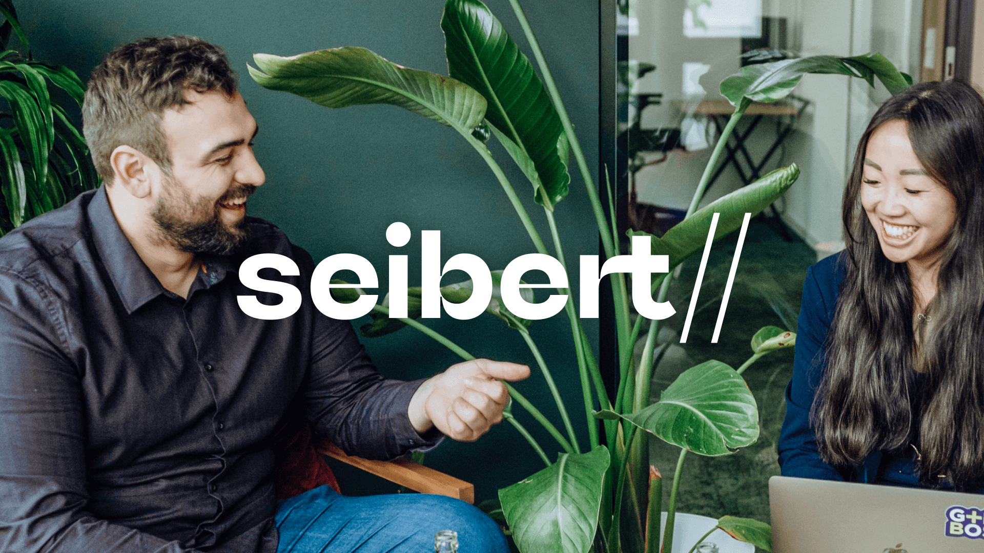 The Seibert Network Has a New Logo - the new Seibert logo with two employees laughing together in the background with a large plant between them