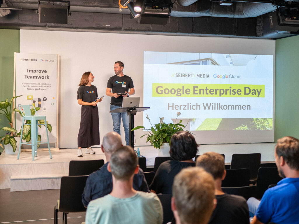 Ein Foto der Bühne vom Google Enterprise Day 2022. Rechts wird die Präsentation an die Wand geworfen. Links davon stehen ein Mann und eine Frau, die beiden Modaratoren des Tages, die sich ansehen. Im Vordergrund sitzen Menschen vor der Bühne.